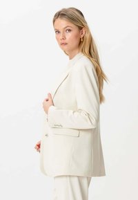 En blond kvinde, iført guld hoop øreringe og en off-white jakkesæt, poserer sidelæns mod en hvid baggrund.
