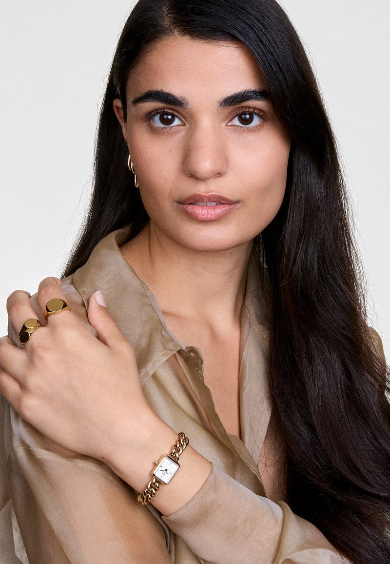 Bracelet en chaîne dorée avec un cadran de montre blanc carré, porté sur une main ; le modèle porte des bagues, a de longs cheveux noirs et une blouse beige transparente.