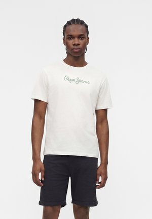 Mężczyzna z warkoczami, ubrany w białą koszulkę "Pepe Jeans" i czarne podwinięte szorty na jednolitym jasnym tle.