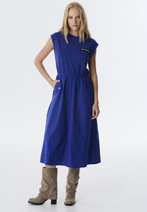 Blauwe, mouwloze jurk met een elastische tailleband, zijzakken en logodetail. Model draagt beige suède enkellaarsjes. Zachte textuur.