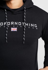 Schwarzer Hoodie mit weißem Text „FOR NOTHING“ und „UNITED KINGDOM“ sowie einem kleinen Union Jack-Patch, schwarze und weiße Kordeln.