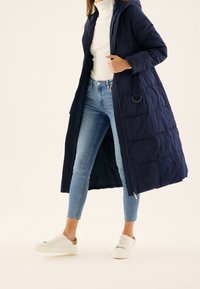 Abrigo largo acolchado en navy con capucha, cremallera frontal y bolsillos laterales, combinado con unos vaqueros ajustados azul claro y zapatillas blancas.