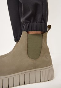 Bottines en cuir taupe avec des panneaux élastiques sur les côtés en vert olive, dotées d'une semelle en caoutchouc grise à nervures et d'une languette pour un enfilage facile.