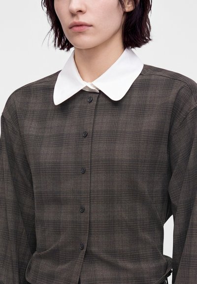 Jeune personne portant une chemise à carreaux foncée boutonnée avec un grand col blanc pointu contrastant sur un fond uni.