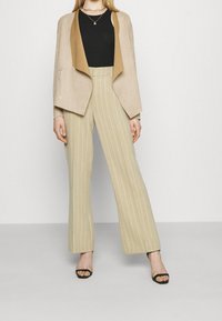 Veste beige perforée avec une insert en marron, associée à un pantalon large rayé crème et des sandales noires à talons.