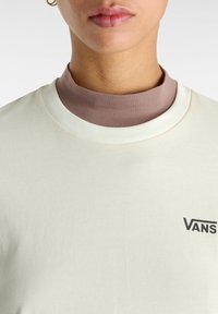 Camiseta de color crema claro con escote redondo, que presenta una capa de cuello simulado marrón debajo. Logotipo negro de "VANS" en el frente. Textura suave.