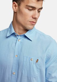 Helles blaues Button-Up-Hemd aus Baumwolle. Mit einer Tasche mit besticktem Design. Klassischer Kragen und orangefarbene Knöpfe. Glatte Textur.