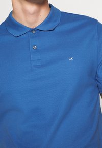 Polo bleu en tissu de coton texturé. Il présente un col à revers, deux boutons et un petit logo "ck" blanc sur la poitrine.