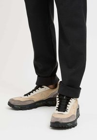 Sneakers con una parte superiore in suede beige e grigia, suola in gomma nera, lacci neri e un dettaglio del logo sul lato. I pantaloni sono arrotolati all'orlo.