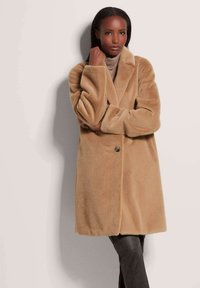 Beige nepbontjas met een notch lapel, sluiting met één knop en brede mouwen, gedragen over een lichte trui en donkere broek.