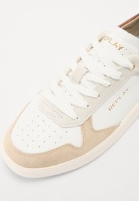 Hvite sneakers med beige semsket detaljer, perforert tå, flate hvite snørebånd og gull "REPLAY"-logo på siden. Gummisåle og teksturert finish.
