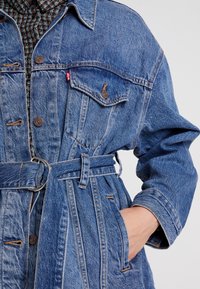 Jeansjacka i ljusblått med långa ärmar, midja med bälte, bröstficka och två sidfickor. Har bruna knappar och sömnadsdetaljer.