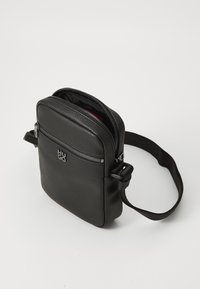 Geantă crossbody din piele neagră cu un finisaj texturat, având un capac cu fermoar, un logo frontal și o curea ajustabilă. Interiorul este căptușit cu o nuanță contrastantă de roșu.