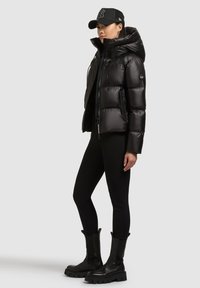 Musta toppatakki, jossa korkea kaulus, vetoketjullinen etuosa ja sivutaskut; yllään mustat leggingsit ja paksupohjaiset mustat saappaat.