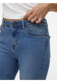 Main ajustant la ceinture d'un jean en denim bleu taille haute, portant un haut blanc et une bague argentée entrelacée à la main droite.