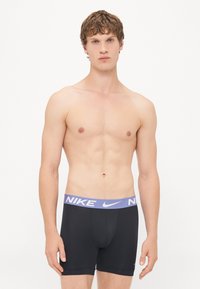 Boxeri negri, cu un brâu violet cu logo-ul Nike, având un design ajustat și o textură netedă, purtați de o persoană cu păr scurt.