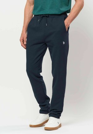 Mann trägt marineblaue Jogginghose mit Kordelzug und weißen Turnschuhen, steht mit einer Hand in der Tasche vor neutralem Hintergrund.