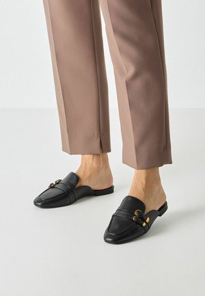 Zwarte leren muiltjes met een puntige neus, voorzien van decoratieve gouden hardware en gestikte accenten. Gecombineerd met lichtbruine taps toelopende broek.