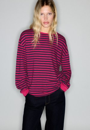 Femme blonde portant une chemise ample rayée horizontale rose et noire à manches longues et un jean bleu foncé sur un fond clair uni.