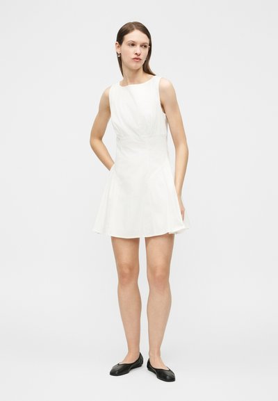 Γυναίκα που στέκεται με τα χέρια στις τσέπες, φοράει αμάνικο λευκό φόρεμα και μαύρα επίπεδα παπούτσια, μπροστά σε απλό φωτεινό φόντο.