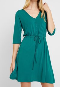 Robe teal en tissu doux avec décolleté en V, manches trois quarts, taille réglable avec cordon de serrage et détail de boutons à l'avant.