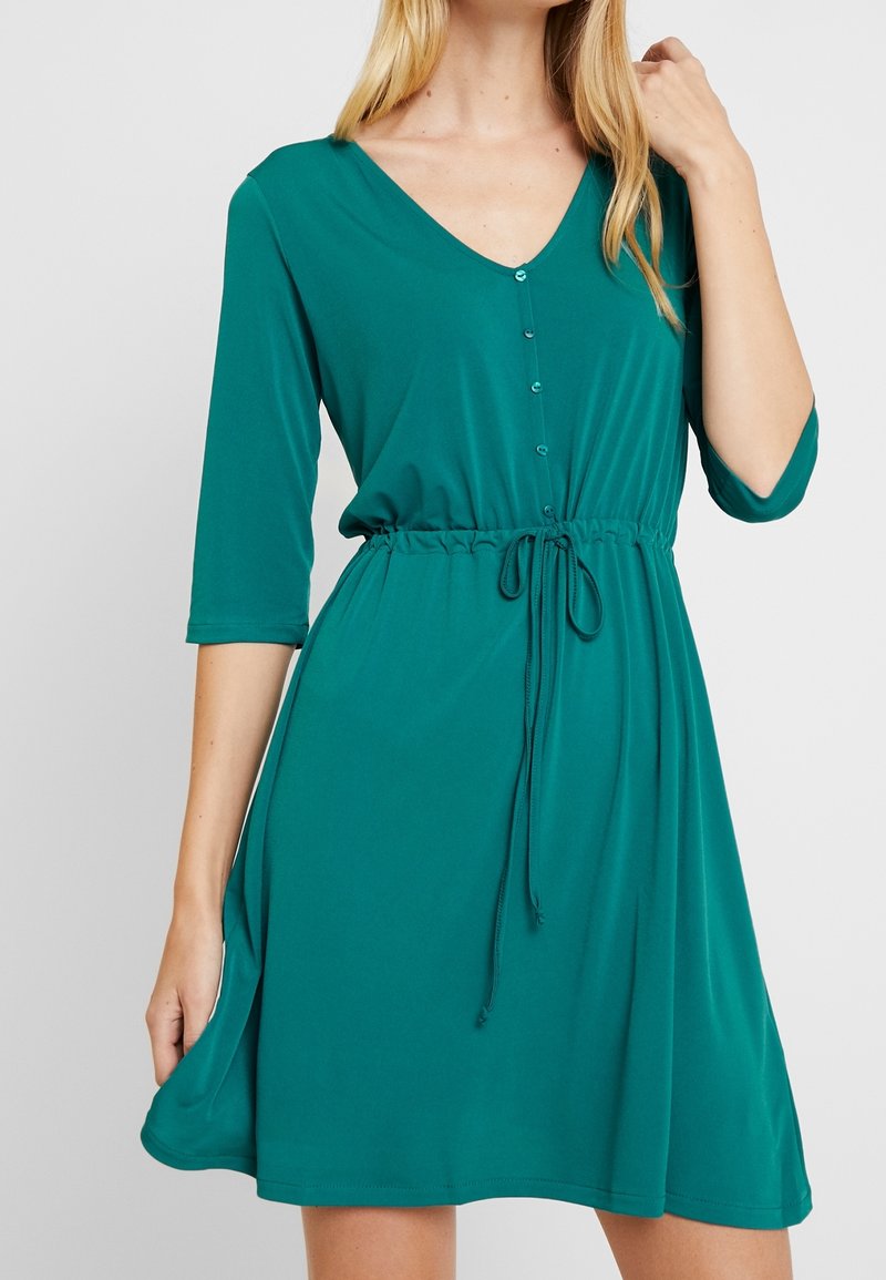 Robe teal en tissu doux avec décolleté en V, manches trois quarts, taille réglable avec cordon de serrage et détail de boutons à l'avant.