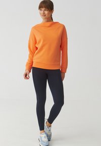 Felpa arancione con un collo alto, abbinata a leggings blu navy e scarpe da ginnastica azzurro chiaro. Il tessuto sembra morbido e elastico.