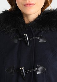 Marinblå ullkappa med svart fuskpäls krage, med toggelspännen med svarta detaljer och texturerade vävda remmar.