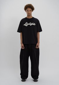 Jauns vīrietis ar cirtainiem matiem, ģērbies melnā, platā "Lowlights" t-kreklā, plašās melnās biksēs un melnās kurpēs, pret vienkāršu pelēku fona.