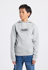 Sudadera con capucha gris con un bolsillo frontal y etiqueta gráfica. Presenta puños y dobladillo acanalados, textura de tela suave. Combinada con jeans azules.