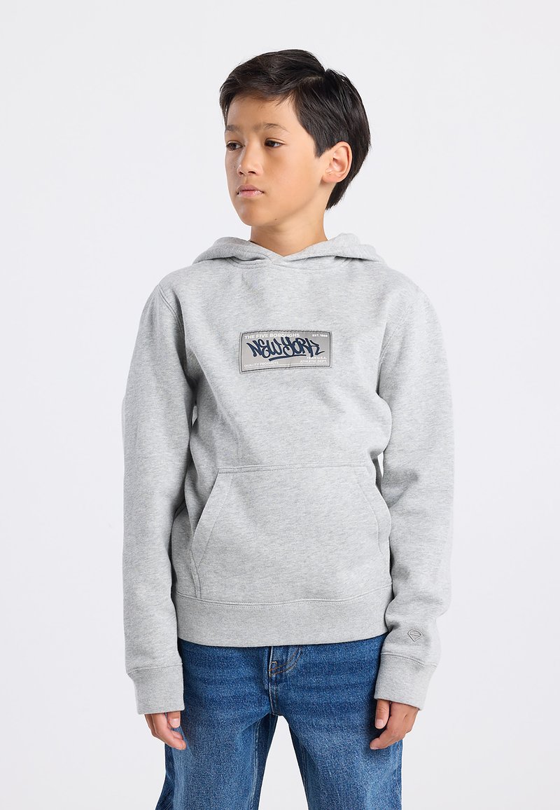 Sudadera con capucha gris con un bolsillo frontal y etiqueta gráfica. Presenta puños y dobladillo acanalados, textura de tela suave. Combinada con jeans azules.