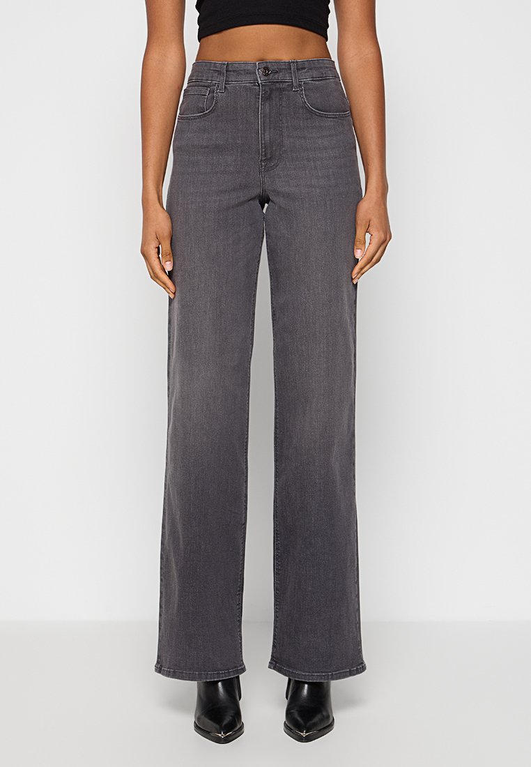 Kvinna klädd i hög midja, vida mörkgrå jeans med framfickor, kombinerat med svarta spetsiga ankelskor och en svart crop top.