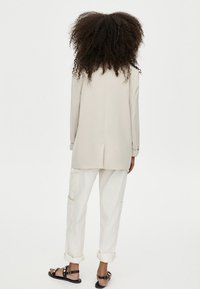 Blazer beige clair à coupe cintrée, porté sur un pantalon cargo blanc ample. Sandales noires complètent la tenue. Cheveux bouclés texturés visibles.