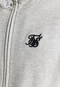 Felpa grigia realizzata in morbido tessuto, con un logo ricamato nero sul petto. Il design prevede un cappuccio con coulisse e una zip frontale.