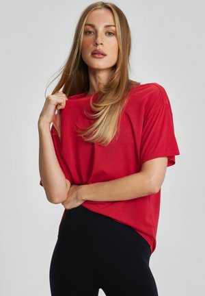 Kobieta o długich blond włosach, ubrana w luźną czerwoną koszulę z krótkim rękawem i czarne spodnie, trzymająca kołnierzyk koszuli jedną ręką.