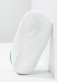 White sneaker sole with a textured surface, featuring a green accent on the heel. Includes circular grip patterns and embossed logo detail.