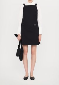 Vestido negro con escote cuadrado y tirantes, combinado con una camisa blanca. Lleva un bolso de mano negro con pliegues. Zapatos planos sencillos completan el look.