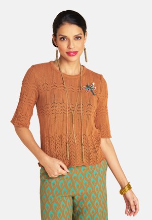 Woman wearing a rust orange knit top with a colorful dragonfly brooch, long gold necklace, patterned green and brown pants, and gold bangles.