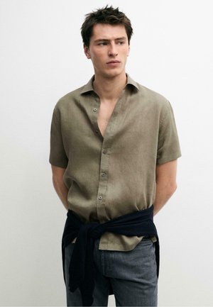 Junger Mann mit kurzen dunklen Haaren, der ein olivgrünes Kurzarmhemd und einen dunklen Pullover um die Taille gebunden trägt, steht vor einer weißen Wand.