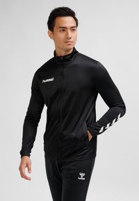 Mężczyzna w czarnej sportowej kurtce na pełny zamek i dopasowanych spodniach z logo hummel, stojący z jedną ręką w kieszeni kurtki, patrzący na bok.