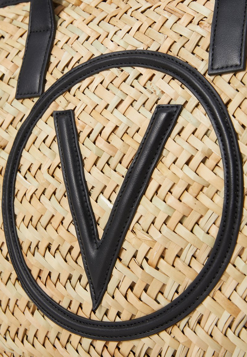 Sac fourre-tout en paille tressée avec des accents en cuir noir. Présente un grand logo circulaire avec un "V" bien en évidence au centre. Surface texturée.