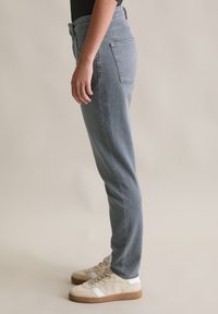 Een persoon in lichtblauwe slim-fit jeans en beige sneakers staat in zijaanzicht tegen een effen lichte achtergrond.