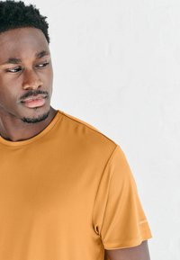 Koszulka z krótkim rękawem odprowadzająca wilgoć, wykonana z musztardowo-żółtej siateczkowej tkaniny, z okrągłym dekoltem i subtelnymi bocznymi akcentami.