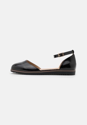 Chaussure plate en cuir verni noir, bout fermé, bride de cheville avec boucle dorée et semelle noire texturée sur fond blanc.