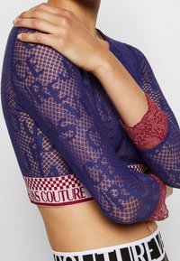 Maglietta a maniche lunghe viola con texture a rete, con dettagli in pizzo sulle maniche e una cintura decorata in rosso e bianco con dettagli di branding.