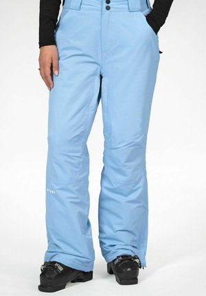 Vaaleansiniset lasketteluhousut mustien laskettelukenkien kanssa, henkilö pukeutuneena, kädet taskuissa, näkyvissä vyötäröstä varpaisiin valkoisella taustalla.