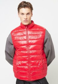 Red puffer vest with a high collar, padded design, and front zipper. The fabric has a glossy finish, contrasting with the gray long-sleeve shirt underneath.