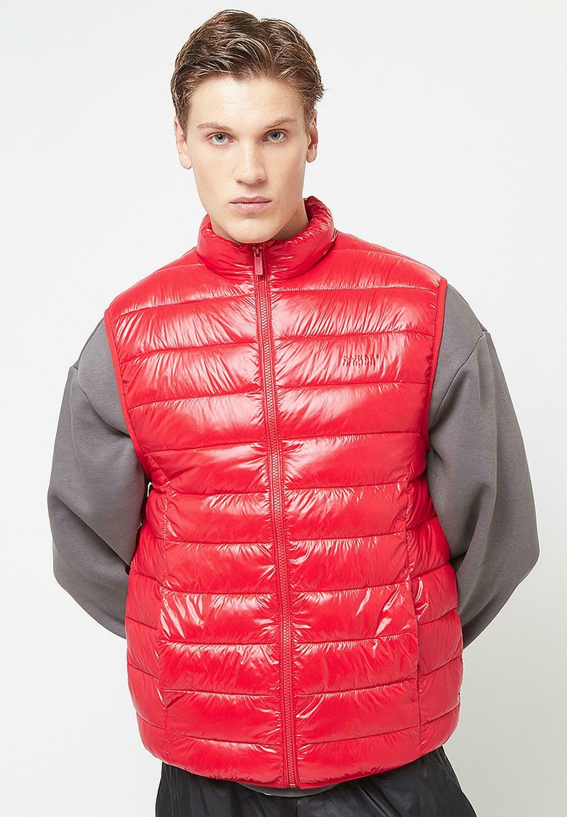 Red puffer vest with a high collar, padded design, and front zipper. The fabric has a glossy finish, contrasting with the gray long-sleeve shirt underneath.