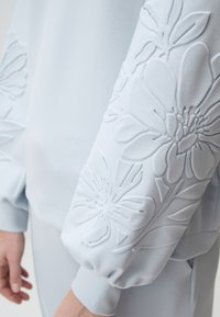 Sweatshirt bleu clair avec des motifs floraux en relief sur les manches, à coupe décontractée et à la texture douce.