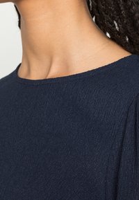 Tessuto strutturato blu navy con scollatura a barca, focalizzandosi sui dettagli delle spalle, enfatizzando la morbidezza e una leggera caduta.